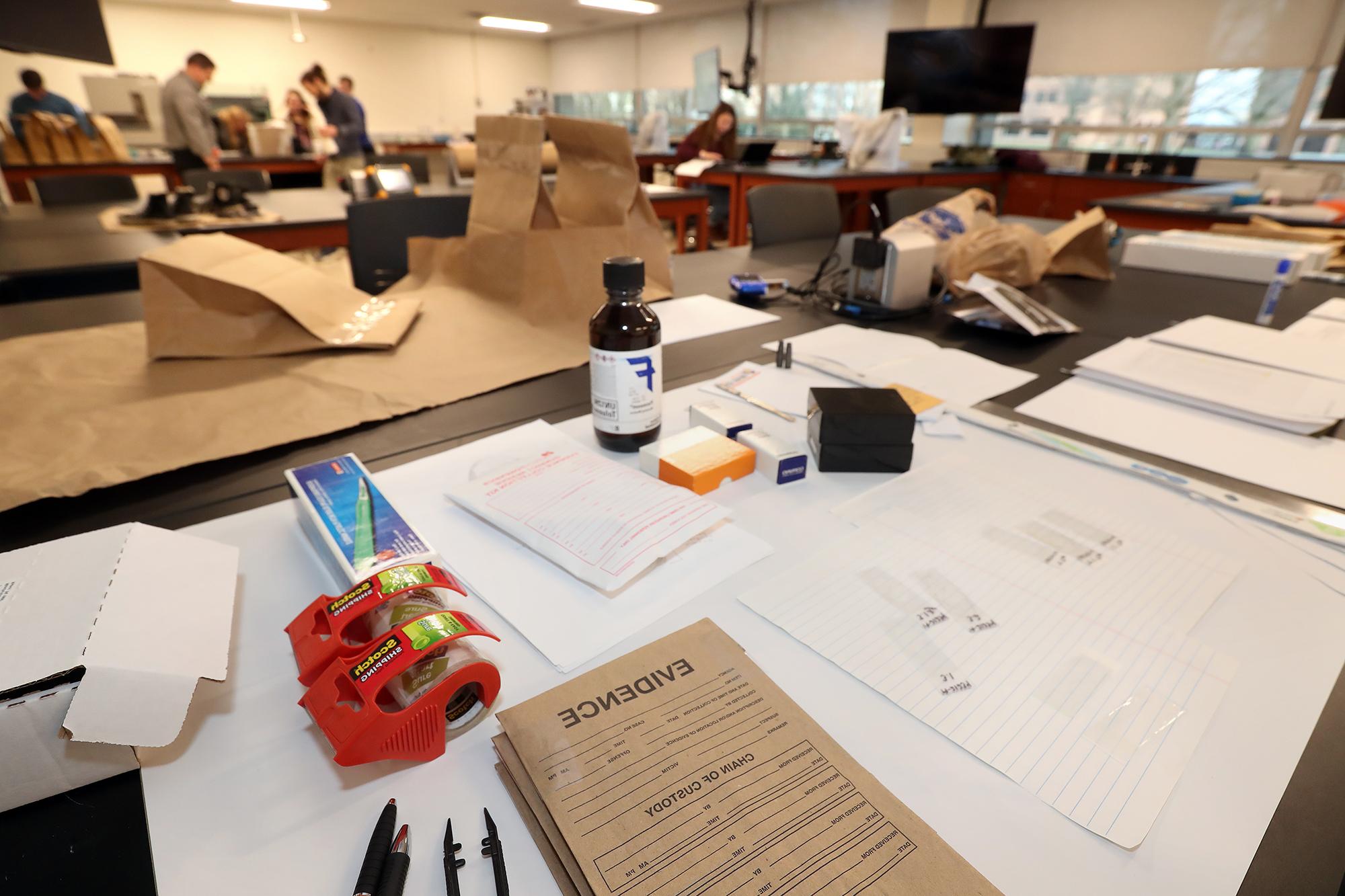 波士顿州立大学法医实验室的一张桌子上有棕色的证据袋, 化学品和其他工具，学生在后台使用，以处理犯罪现场的证据.